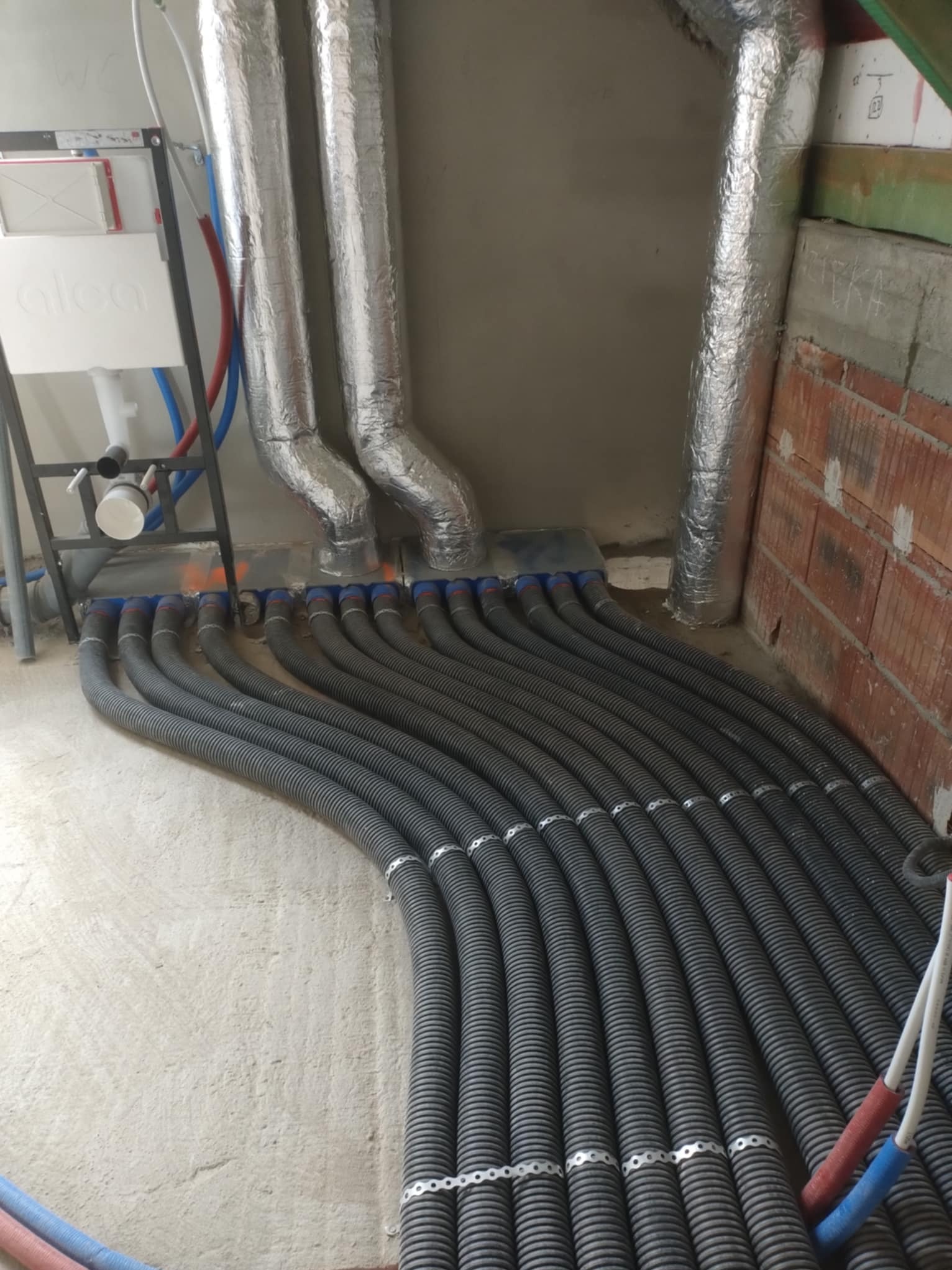 Szereg czarnych, karbowanych rur wentylacyjnych podłączonych do rozdzielacza z niebieskimi końcówkami, z widocznymi izolowanymi przewodami oraz aluminiowymi kanałami wentylacyjnymi w surowym...