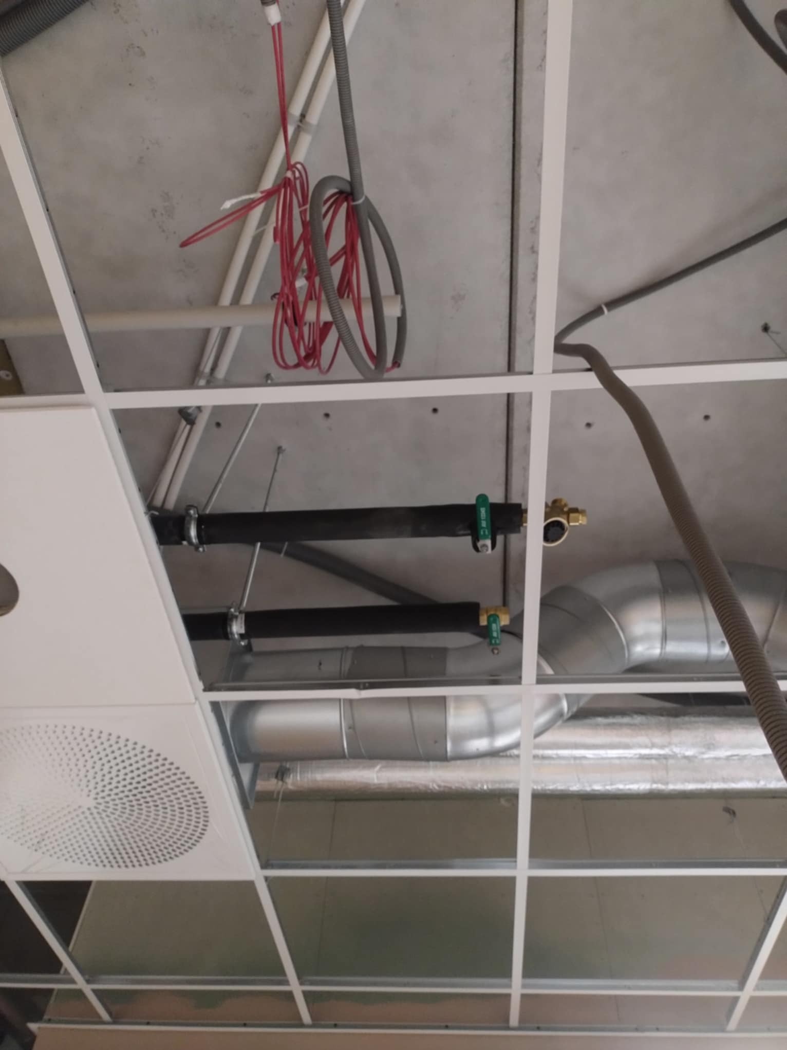Widok instalacji pod sufitem podwieszanym z widocznymi przewodami wentylacyjnymi, rurami w izolacji, kablami oraz kratką wentylacyjną.