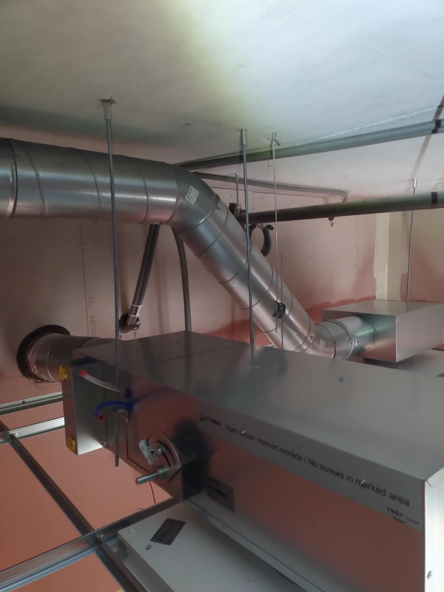 Widok na system wentylacyjny z metalowymi rurami i urządzeniem TROX, zamontowany pod sufitem w pomieszczeniu z instalacją podwieszanego sufitu.