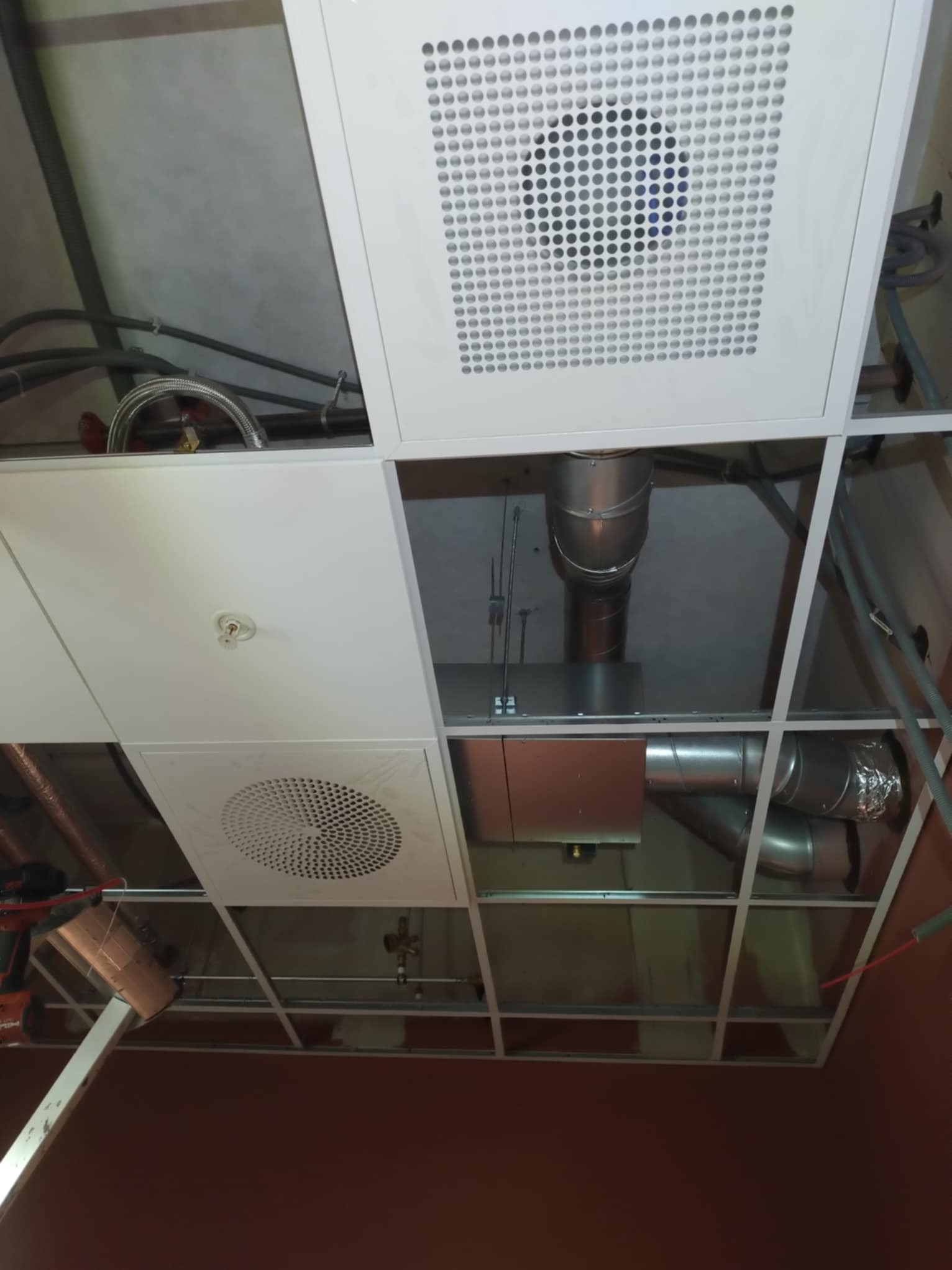 Widok z dołu na podwieszany sufit z zamontowanymi elementami instalacji wentylacyjnej, w tym kratki wentylacyjne z okrągłymi otworami, kanały wentylacyjne oraz instalacja przeciwpożarowa.