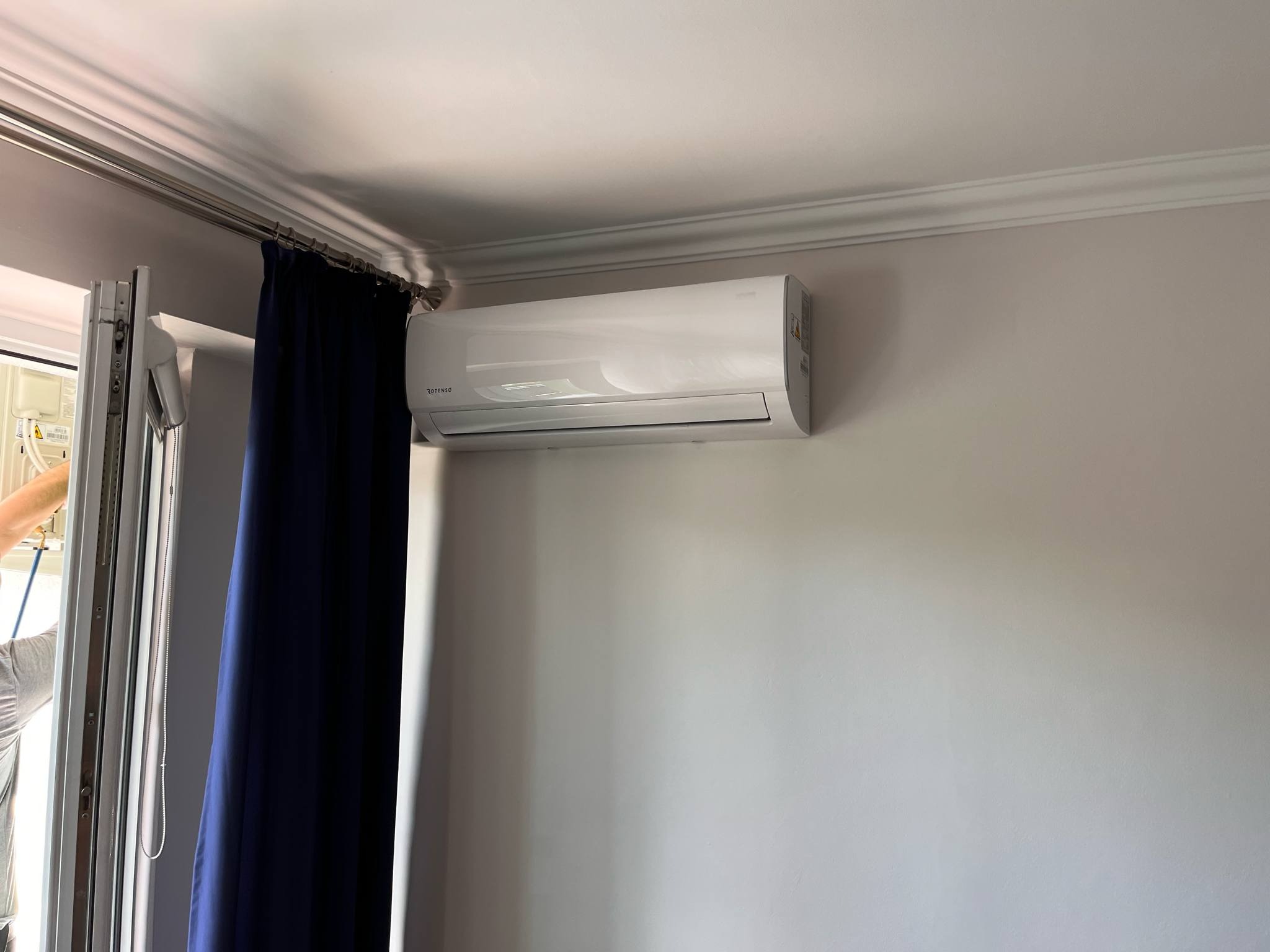 Biała jednostka klimatyzacyjna marki Rotenso zamontowana na szarej ścianie w pokoju z dekoracyjną listwą przysufitową i zasłoniętym oknem.
