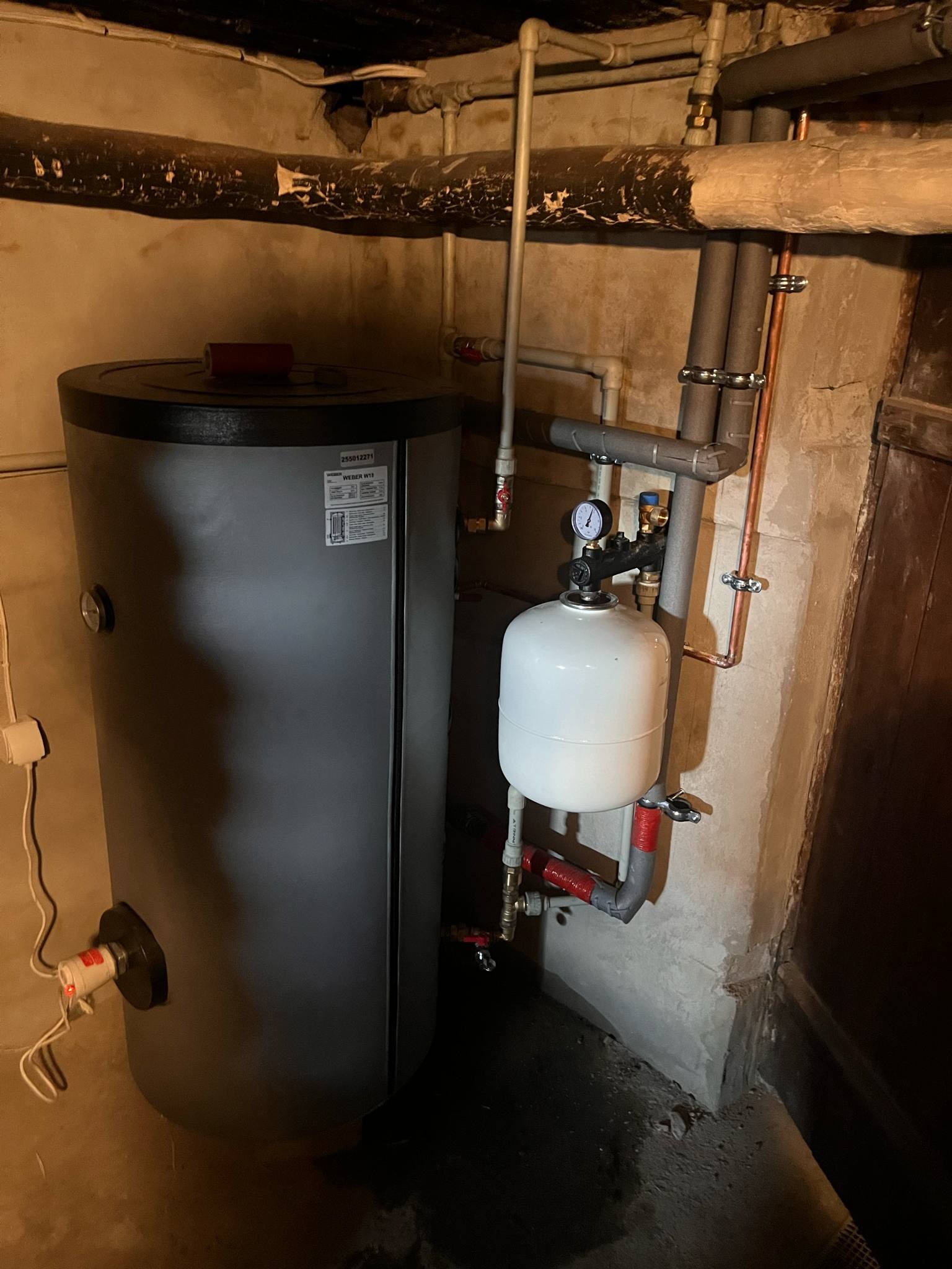 Szary bojler Weber W18 z podłączonym białym zbiornikiem przeponowym i instalacją hydrauliczną w piwnicy otynkowanej jasną zaprawą.