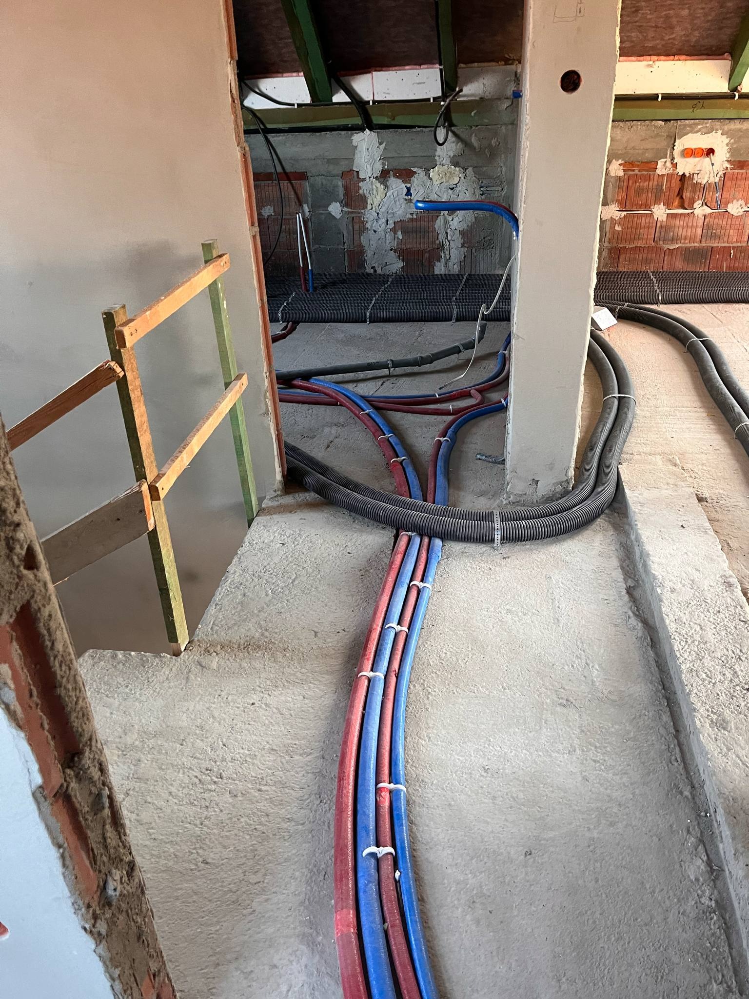 Wnętrze w trakcie prac instalacyjnych: wiązka rur PEX czerwono-niebieskich wychodząca z podłogi, szare rury karbowane na posadzce, surowy beton, fragment ściany z cegły i drewniany stelaż.