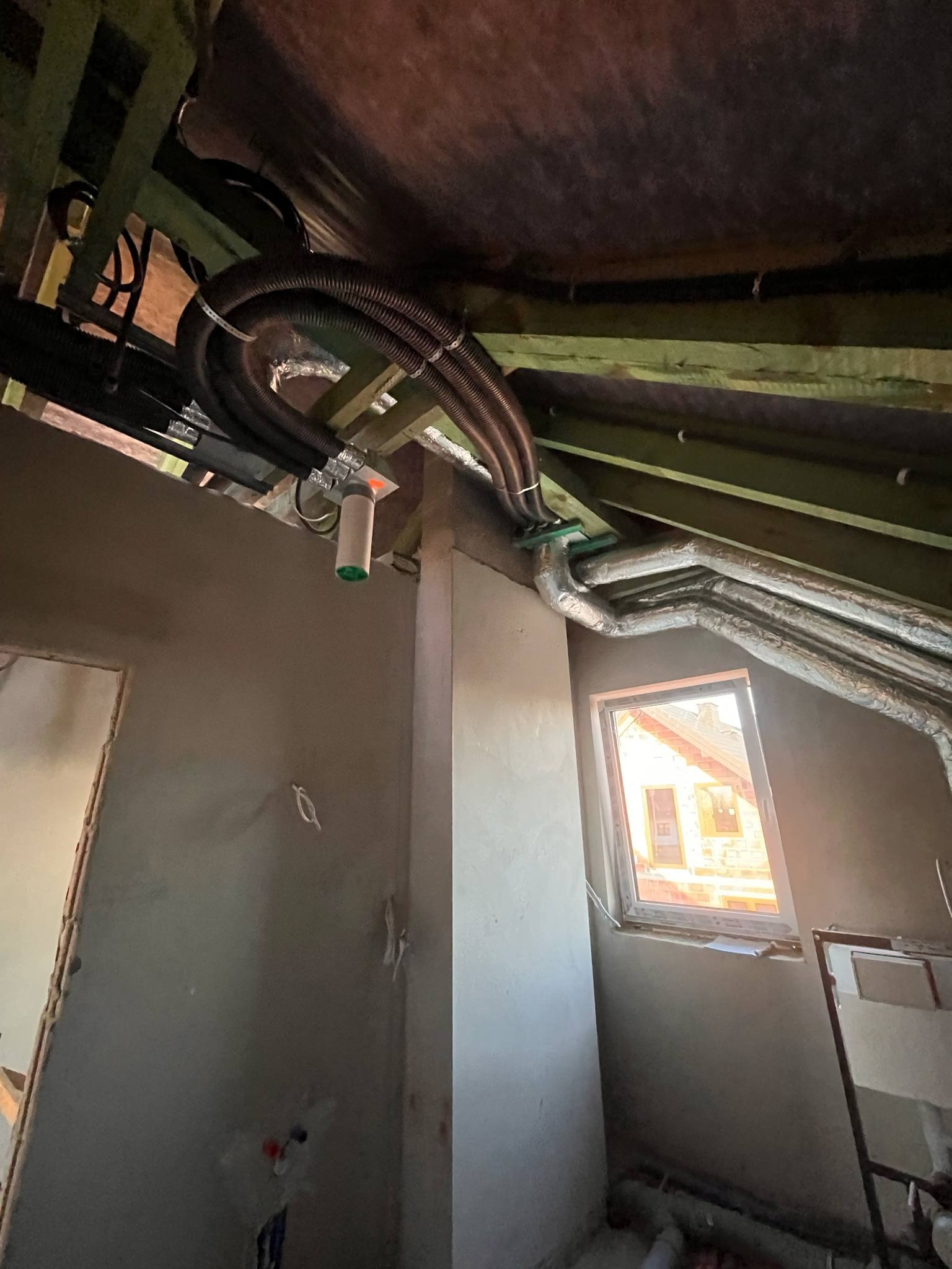Wnętrze poddasza w trakcie prac wykończeniowych; widoczne czarne i srebrne rury wentylacyjne pod stropem, fragment ściany w surowym tynku i okno z widokiem na sąsiedni dom.