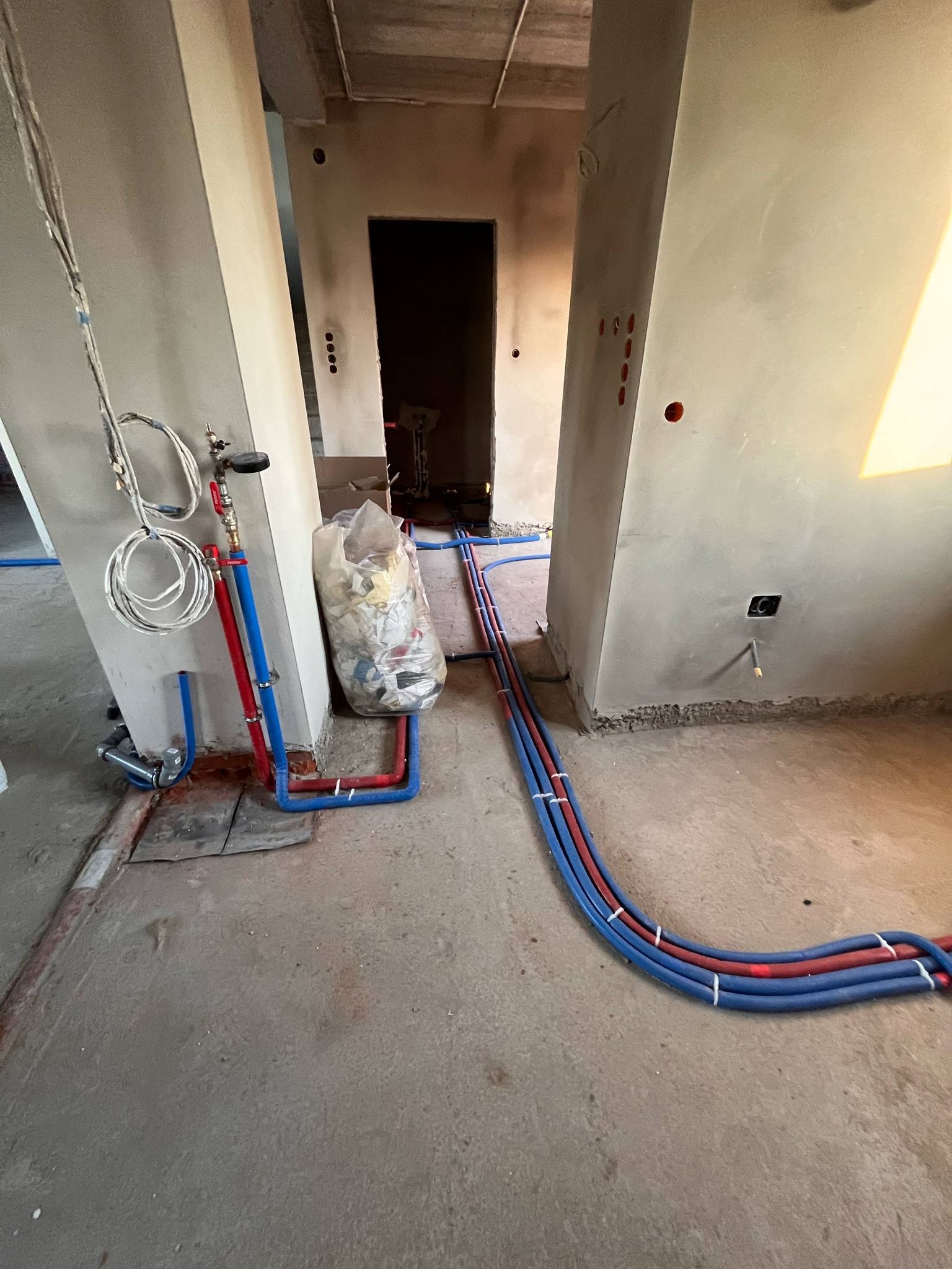 Surowe wnętrze w trakcie remontu z widocznymi, częściowo poprowadzonymi rurami instalacji wodnej w kolorach niebieskim i czerwonym, ułożonymi na surowym betonie, oraz workiem odpadów budowlanych...
