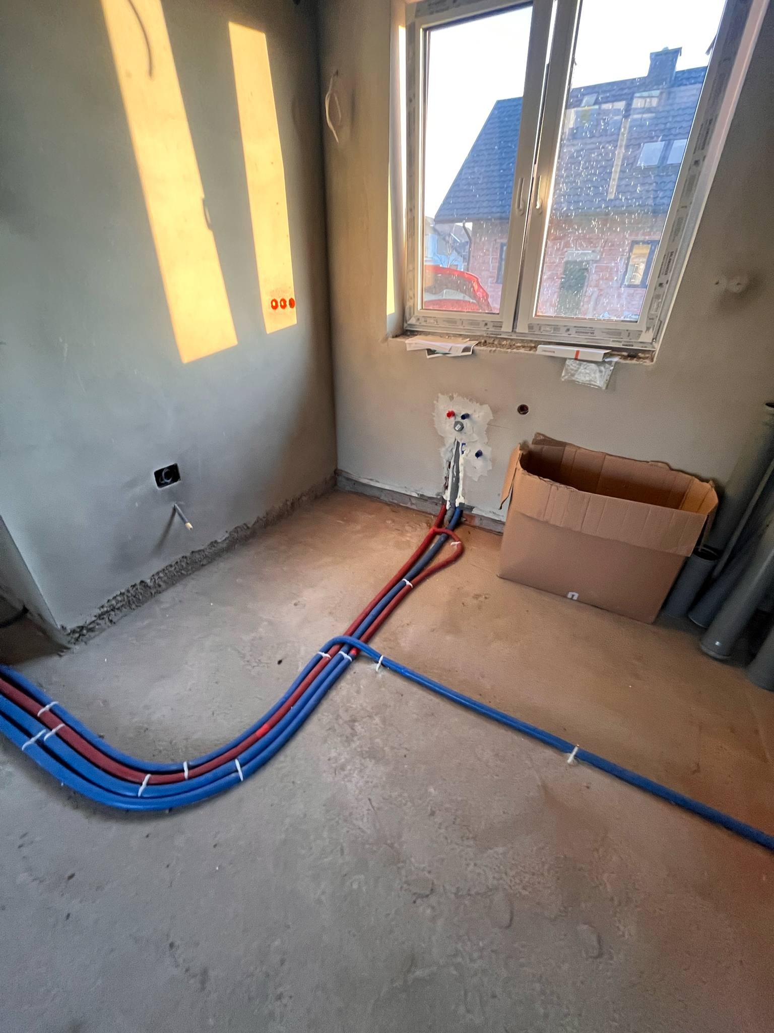 Wnętrze w stanie surowym z poprowadzonymi rurami instalacji wodnej (czerwone i niebieskie) przy ścianie i oknie, widoczne ślady po kuciu i kartonowe pudło.