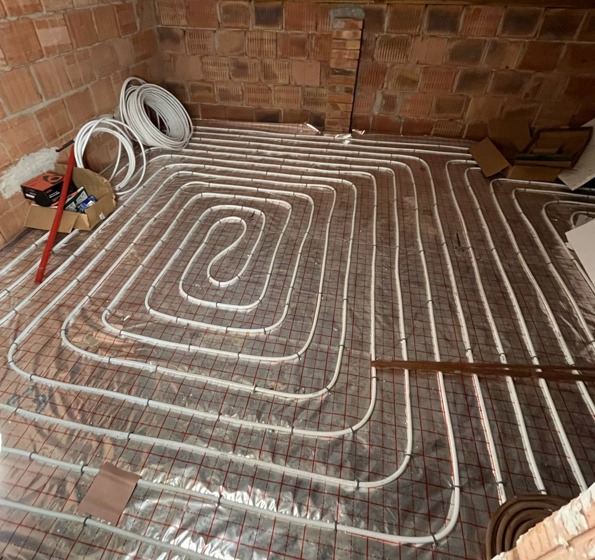 Instalacja ogrzewania podłogowego z białych rurek ułożonych w pętle na srebrnej folii z czerwoną siatką na tle ceglanej ściany i podłogi w trakcie montażu.