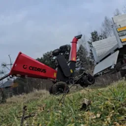 Czerwona rębarka do gałęzi marki Cedrus z podłączoną przyczepą, widoczna na trawiastym terenie z resztkami drewna, pochmurne niebo w tle.