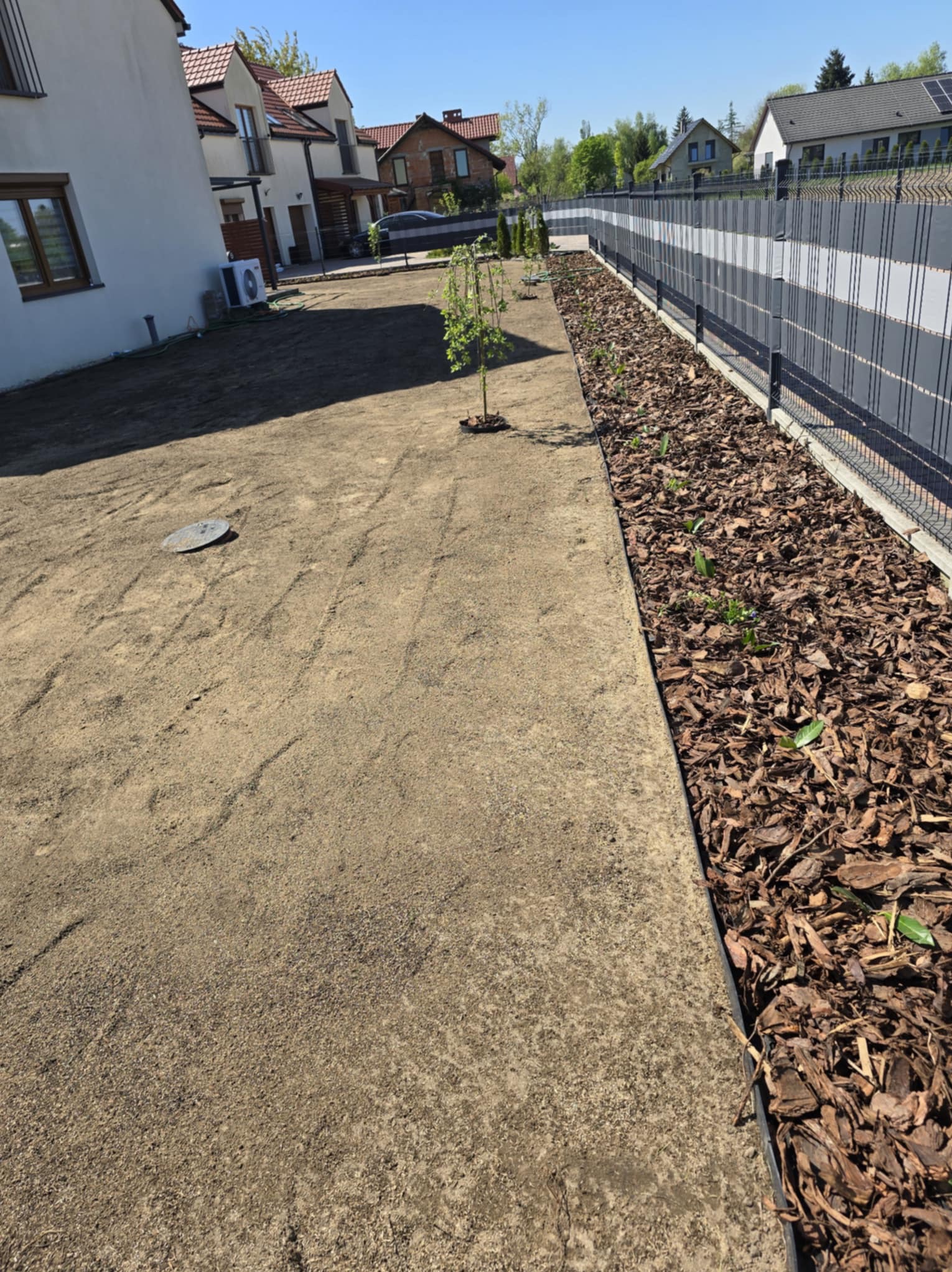 Świeżo założony trawnik z młodymi drzewkami i rabatą wyłożoną korą, oddzieloną od trawnika obrzeżem, wzdłuż nowoczesnego ogrodzenia panelowego.