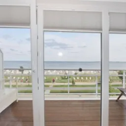 Widok z balkonu na plażę z drewnianą podłogą, białymi zasłonami i roletami w tle.