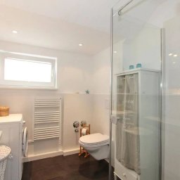 M&K Cleaning Service Klaudia Drachynicz - Jasna łazienka z białą ceramiką, grzejnikiem drabinkowym i kabiną prysznicową ze szkła, drewniany akcent dekoracyjny na ścianie, kosze wiklinowe na pralce.