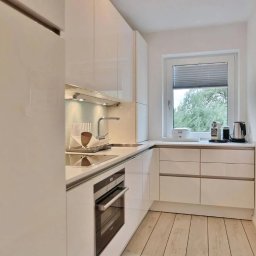 M&K Cleaning Service Klaudia Drachynicz - Błyszcząca, biała kuchnia z drewnianą podłogą, widok na zieleń za oknem, minimalistyczny design, czajnik i ekspres do kawy na blacie.