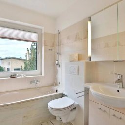 M&K Cleaning Service Klaudia Drachynicz - Jasna łazienka z wanną, toaletą i umywalką, okno z roletą w tle budynku mieszkalnego.