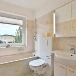 Jasna łazienka z wanną, toaletą i umywalką, okno z roletą w tle budynku mieszkalnego.