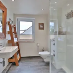 Jasna łazienka z drewnianą ramą lustra o nieregularnej fakturze, drabina z ręcznikami, kabina prysznicowa ze szklanymi drzwiami i panelami oraz szara podłoga z płytek.