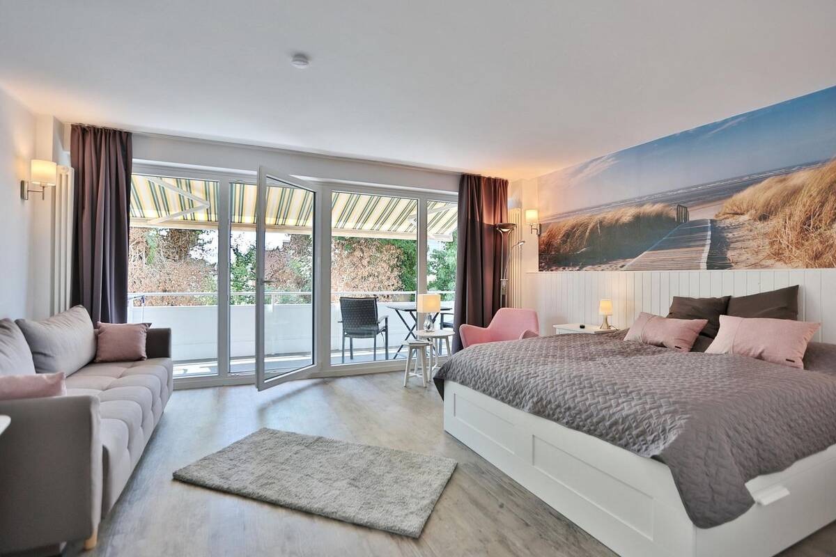 Jasne, przestronne wnętrze apartamentu z balkonem: szary narożnik, łóżko z szarą narzutą i różowymi poduszkami, fototapeta z motywem plaży, drewniana podłoga.