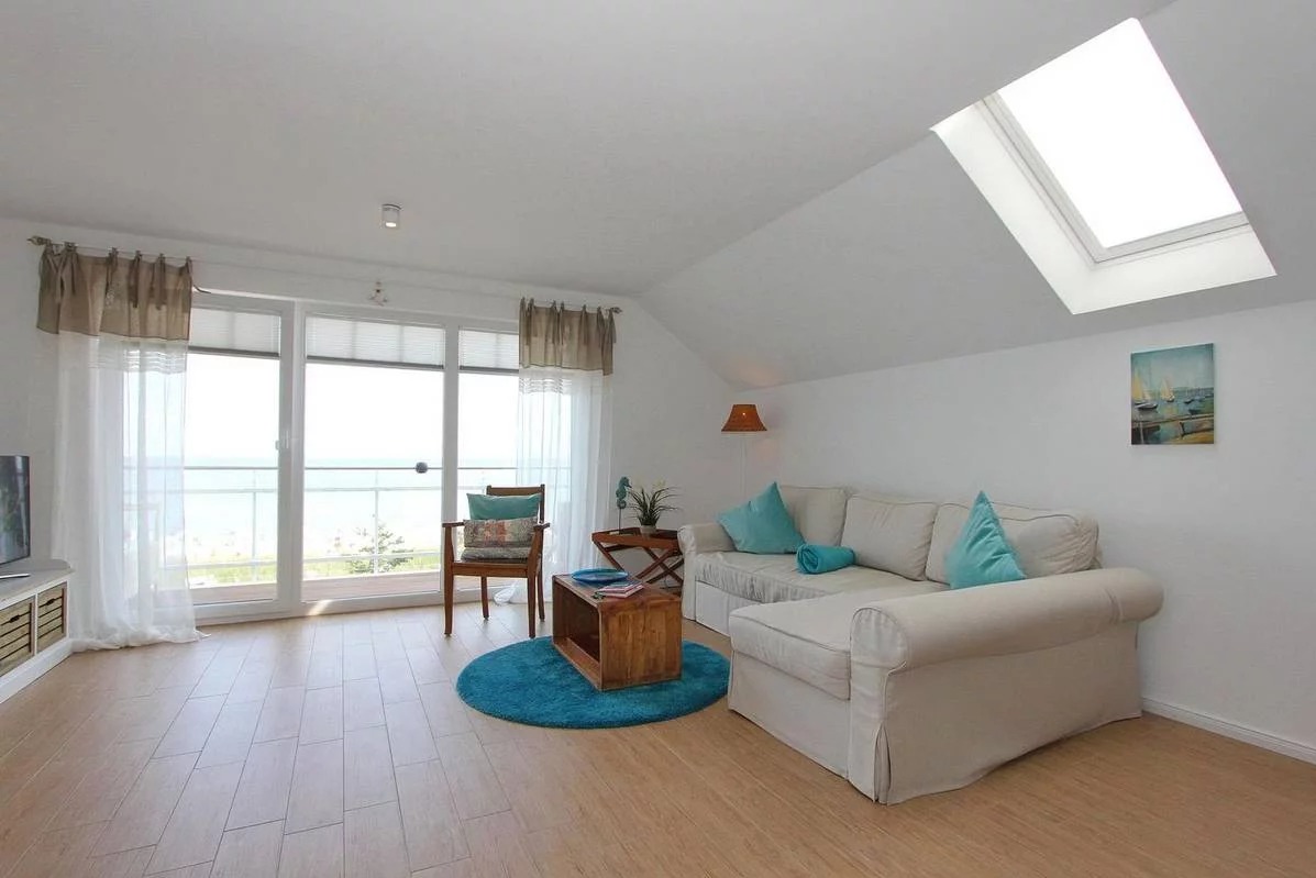 Jasny salon w stylu nadmorskim z drewnianą podłogą, białymi ścianami, sofą z turkusowymi poduszkami, widokiem na morze z balkonu i oknem dachowym.