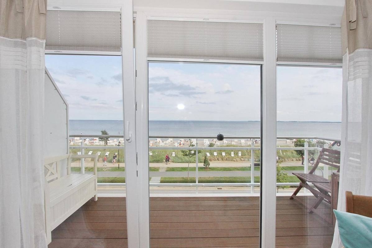 Widok z balkonu na plażę z drewnianą podłogą, białymi zasłonami i roletami w tle.