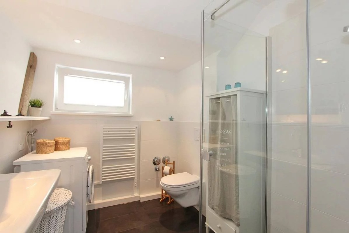 Jasna łazienka z białą ceramiką, grzejnikiem drabinkowym i kabiną prysznicową ze szkła, drewniany akcent dekoracyjny na ścianie, kosze wiklinowe na pralce.