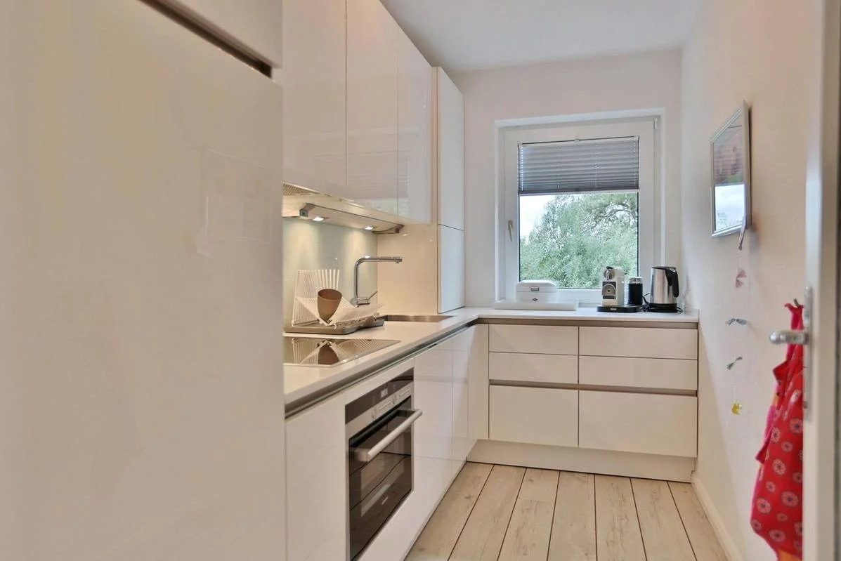 Błyszcząca, biała kuchnia z drewnianą podłogą, widok na zieleń za oknem, minimalistyczny design, czajnik i ekspres do kawy na blacie.