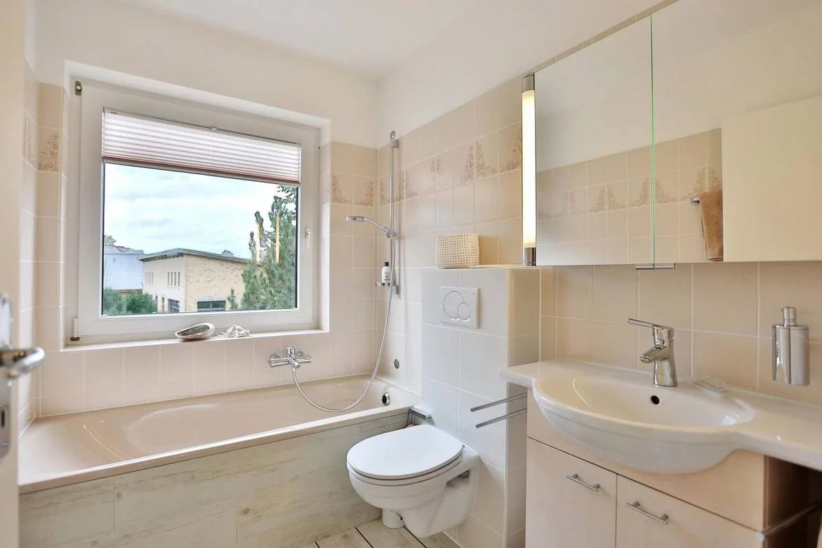 Jasna łazienka z wanną, toaletą i umywalką, okno z roletą w tle budynku mieszkalnego.