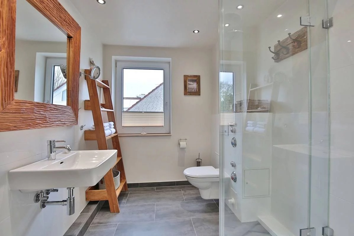 Jasna łazienka z drewnianą ramą lustra o nieregularnej fakturze, drabina z ręcznikami, kabina prysznicowa ze szklanymi drzwiami i panelami oraz szara podłoga z płytek.