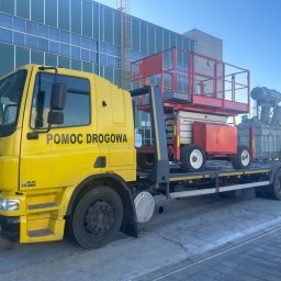 Firma Handlowo Usługowa Jarosław JANIK - Żółta laweta z napisem 'Pomoc Drogowa' przewozi czerwony podnośnik koszowy na tle hali przemysłowej i transformatorów.