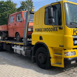 Firma Handlowo Usługowa Jarosław JANIK - Żółta laweta DAF z napisem 'Pomoc Drogowa' przewozi pomarańczowy samochód dostawczy na platformie. Widok z boku, na pierwszym planie chodnik z kostki brukowej.