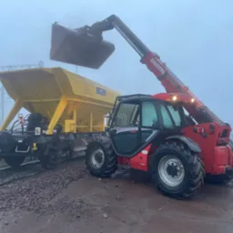 Czerwona ładowarka teleskopowa Manitou z łyżką podnosi materiał do żółtego wagonu kolejowego w mglisty dzień na torach.