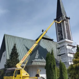 Żółty pojazd z wysięgnikiem podnosi pracownika do wieży kościoła, obok rusztowanie, błękitne niebo w tle.