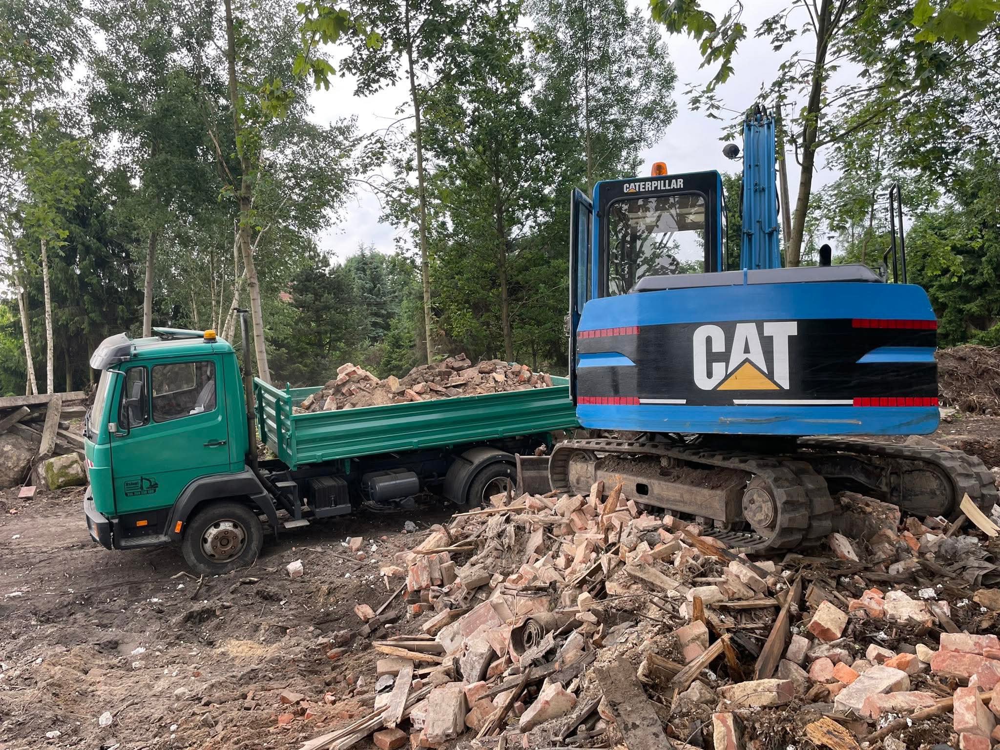 Zielona ciężarówka z ładunkiem gruzu i niebieska koparka CAT na terenie rozbiórki, otoczone drzewami.