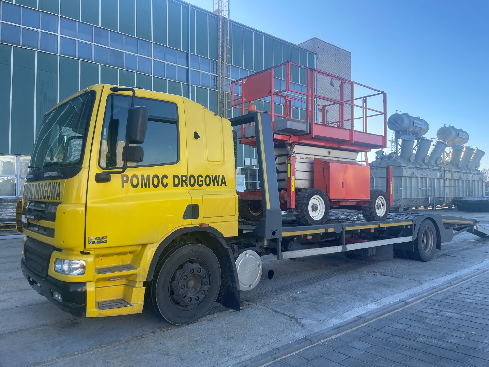 Żółta laweta z napisem 'Pomoc Drogowa' przewozi czerwony podnośnik koszowy na tle hali przemysłowej i transformatorów.