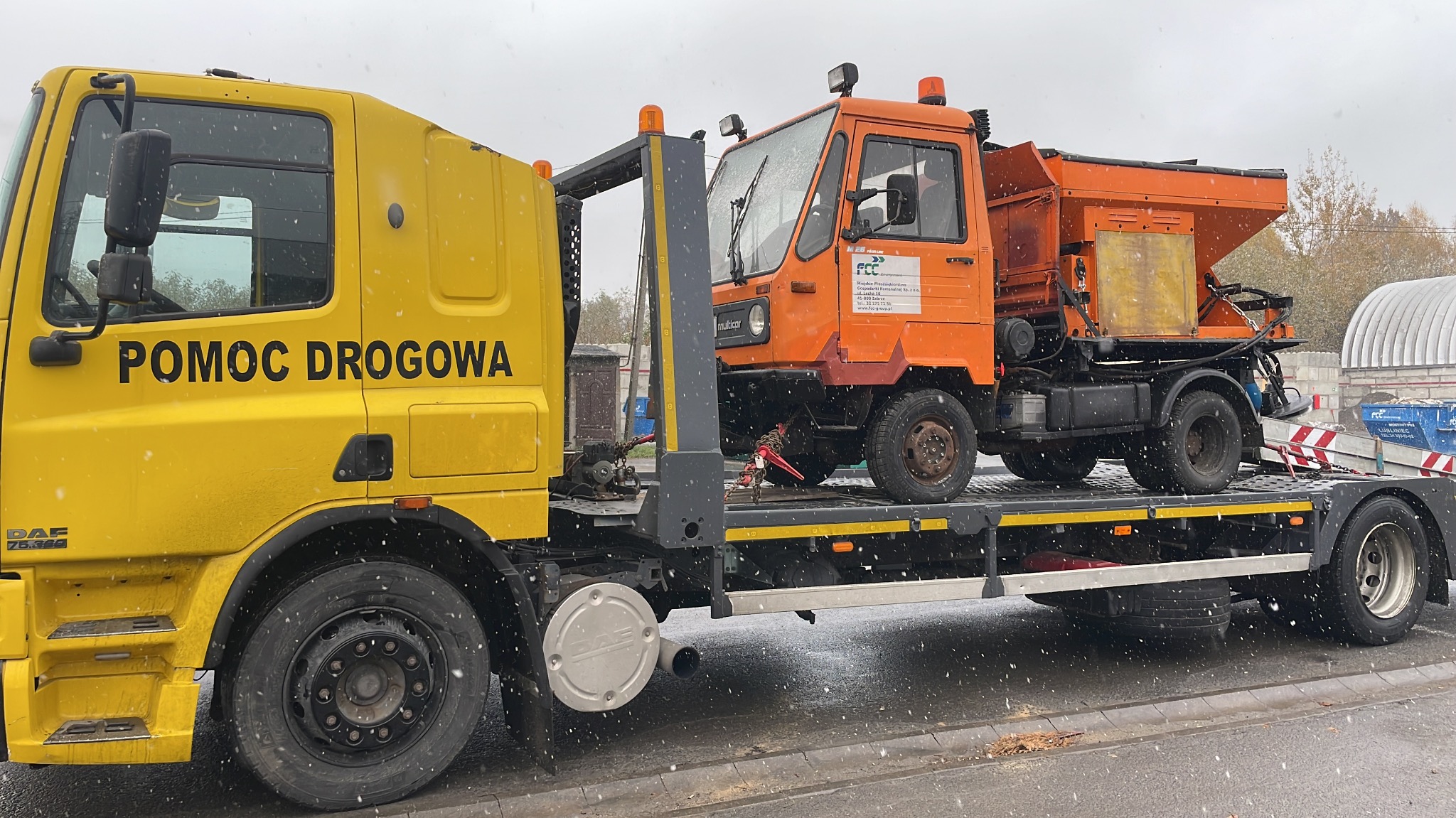 Żółta laweta z napisem 'POMOC DROGOWA' przewozi pomarańczowy pojazd Multicar z posypywarką do soli, widoczne logo FCC.