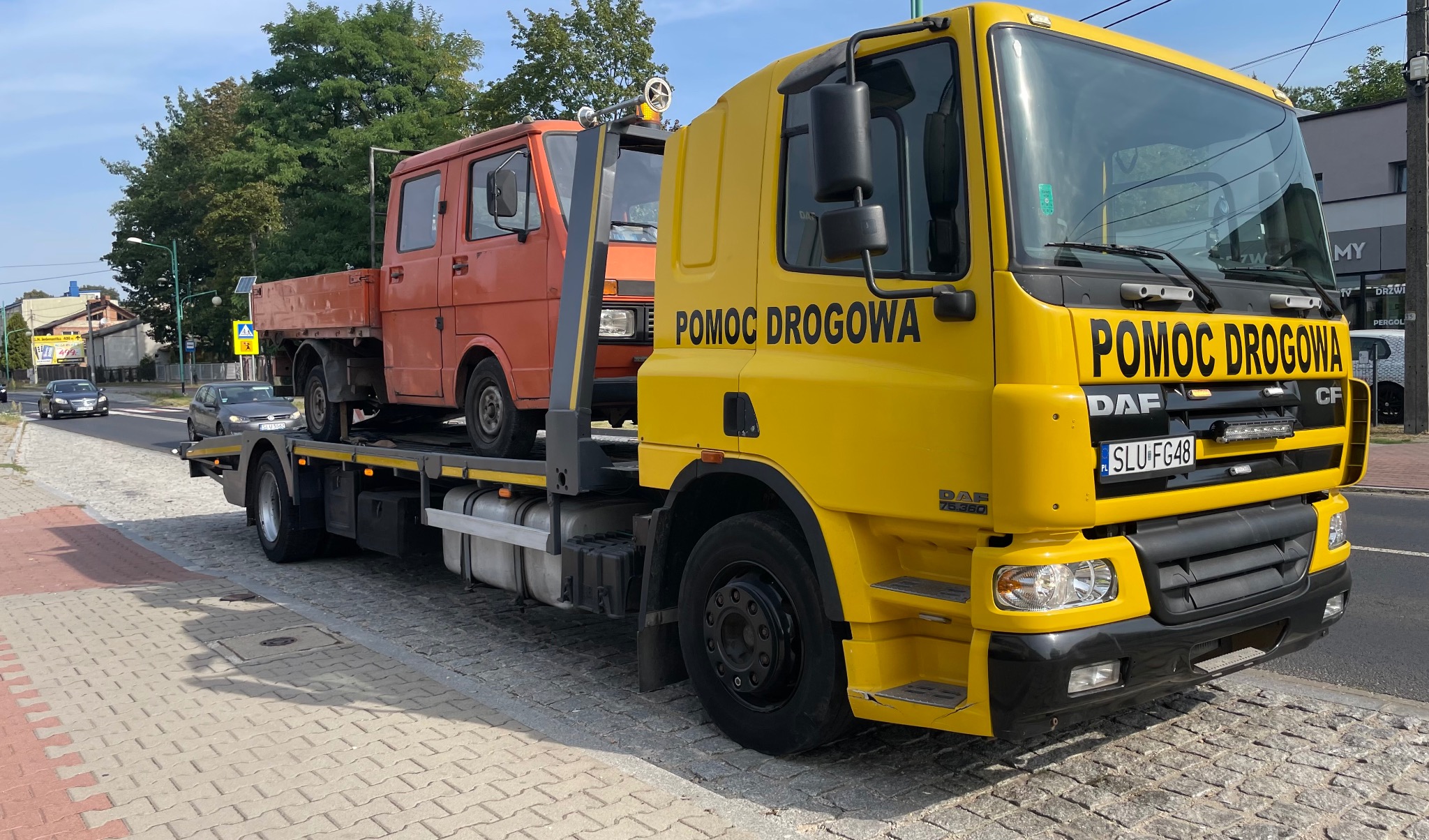 Żółta laweta DAF z napisem 'Pomoc Drogowa' przewozi pomarańczowy samochód dostawczy na platformie. Widok z boku, na pierwszym planie chodnik z kostki brukowej.