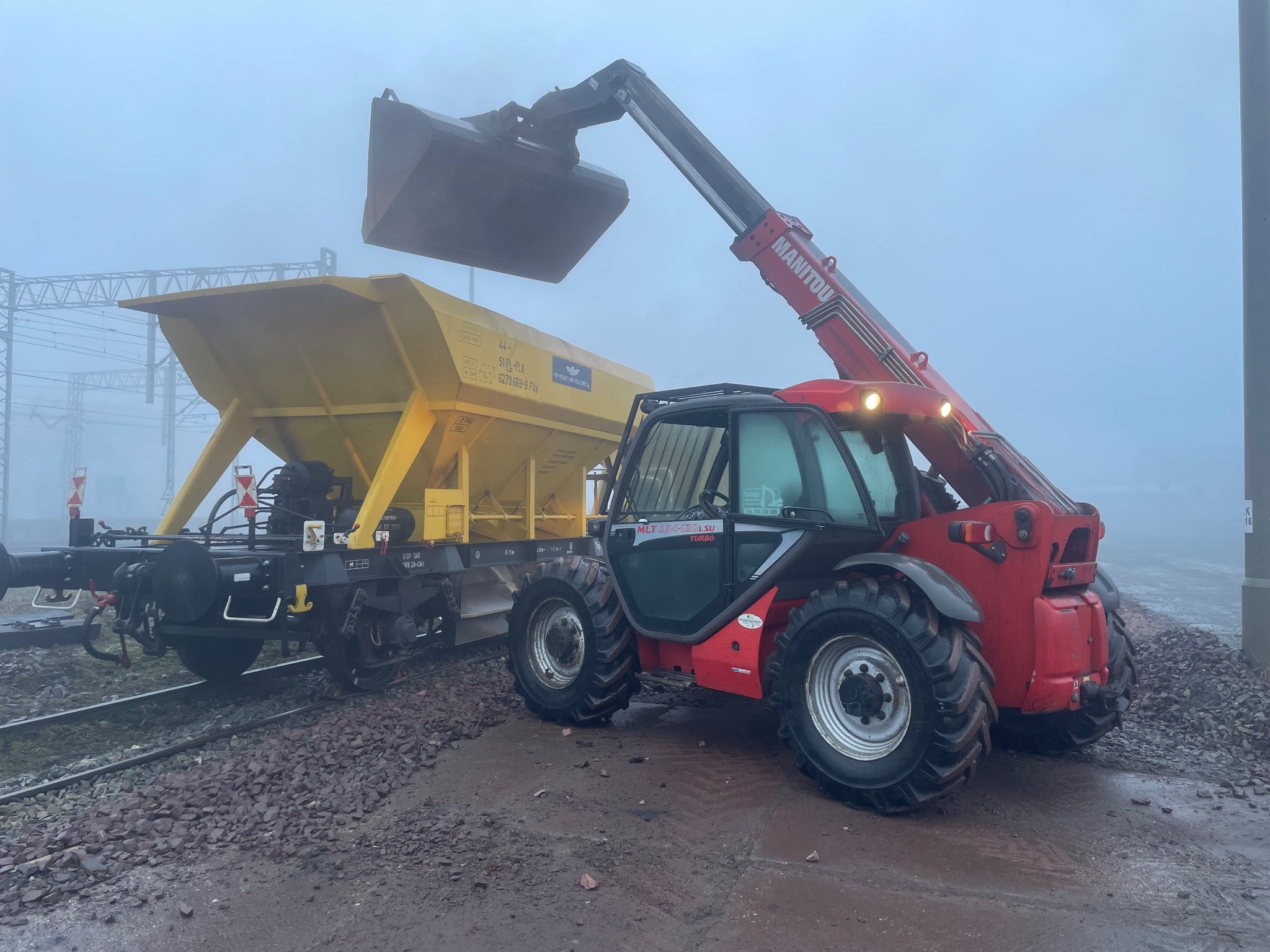 Czerwona ładowarka teleskopowa Manitou z łyżką podnosi materiał do żółtego wagonu kolejowego w mglisty dzień na torach.