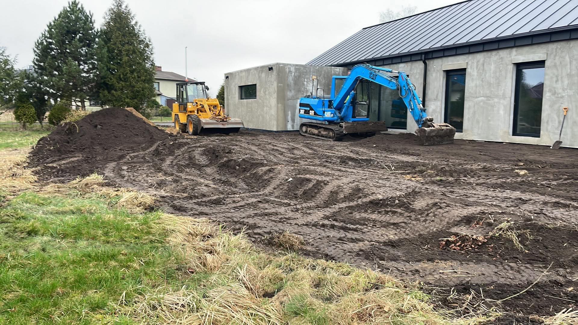 Niebieska mini koparka CAT i żółta ładowarka na placu budowy, wyrównywanie terenu przed nowoczesnym domem z szarą elewacją i czarnymi oknami.