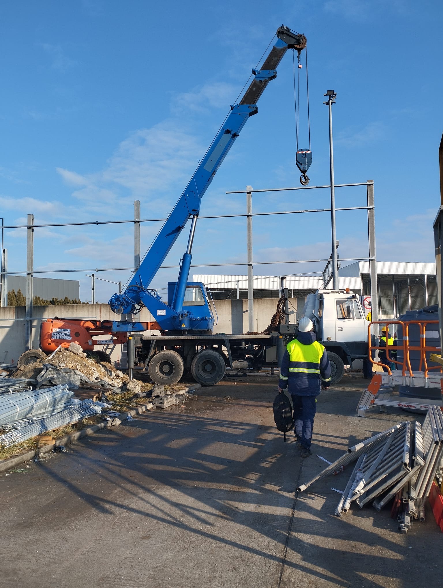 Żuraw samochodowy z wysięgnikiem podniesionym do pionu, w tle konstrukcja stalowa budynku w trakcie montażu, pracownik w odblaskowej kamizelce idzie w stronę żurawia.