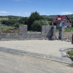 Michał Kachnic - Budowa ogrodzenia z szarej kostki betonowej i układanie kostki brukowej na podjeździe, w tle dom z czerwonym dachem i zieleń. Widoczny pracownik w trakcie pracy.