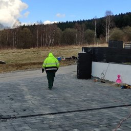 Michał Kachnic - Ułożona szara kostka brukowa na podjeździe, widoczny fragment ogrodzenia z czarnych paneli, pracownik w odblaskowej kurtce. W tle zalesione wzgórza i błękitne niebo.