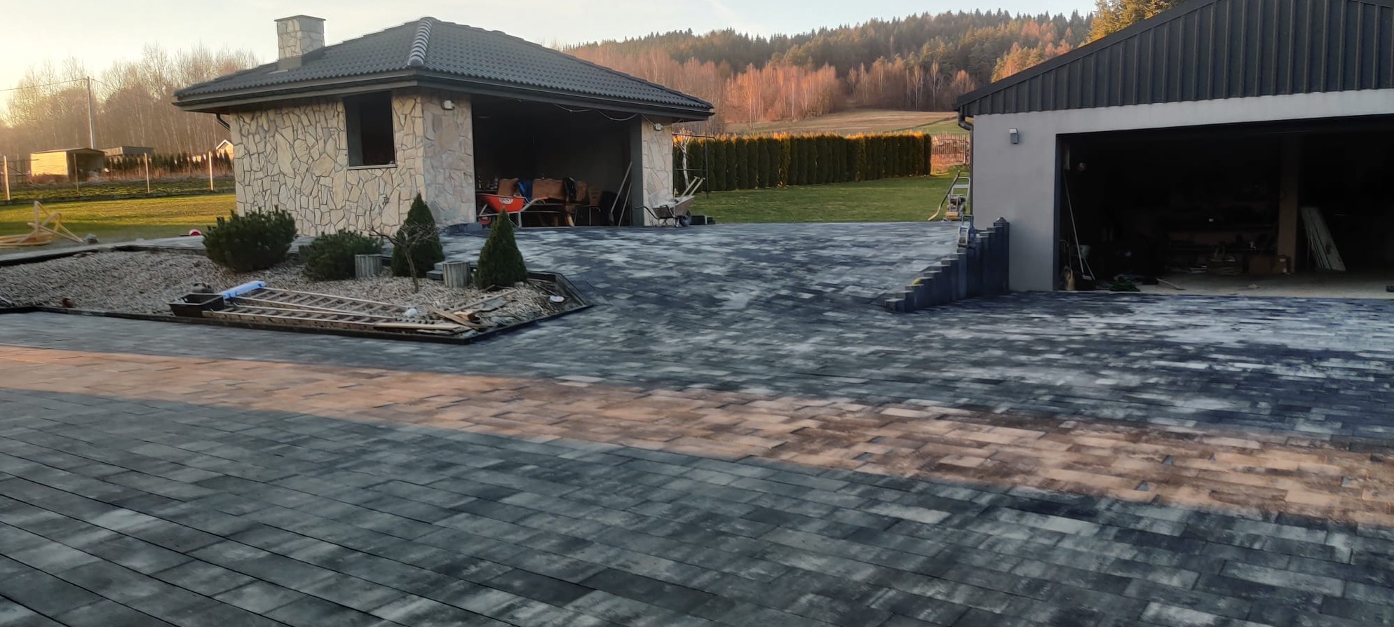 Ułożona kostka brukowa w odcieniach szarości prowadząca do garażu i budynku gospodarczego z kamienia. Widoczne schody z kostki i elementy ogrodu.