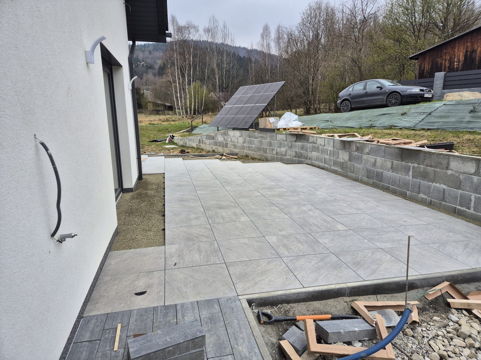 Układanie szarych płyt tarasowych na zewnątrz domu z panelami słonecznymi w tle. Widoczne narzędzia i materiały budowlane. Częściowo ukończona realizacja.
