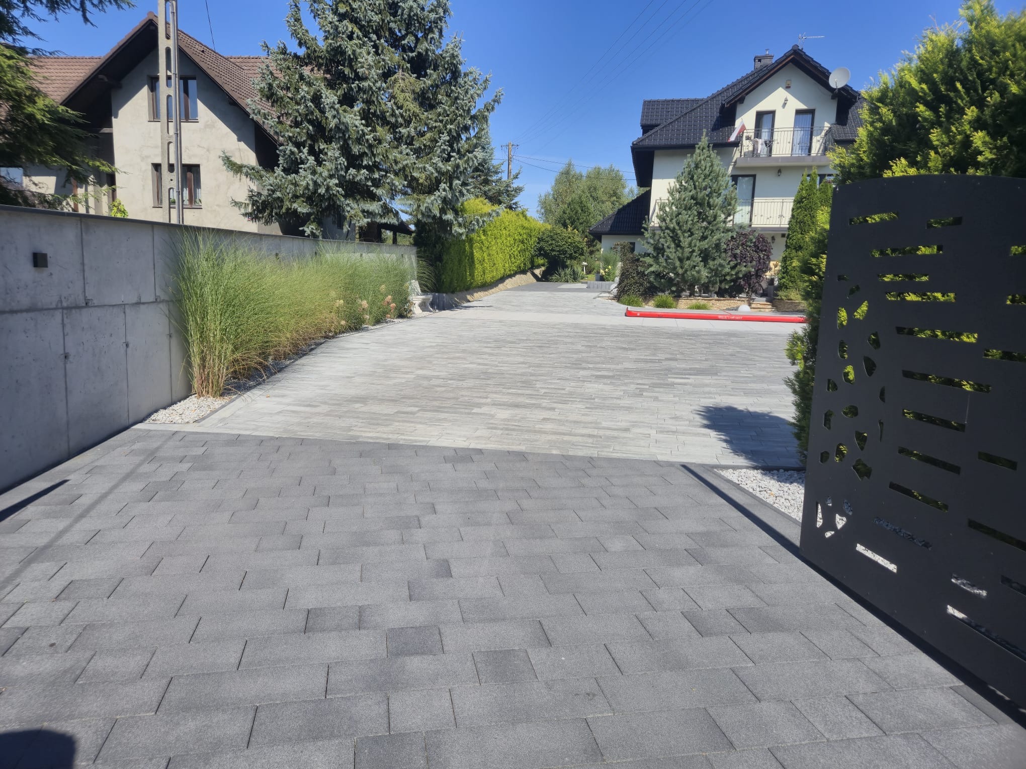 Ułożona szara kostka brukowa na podjeździe, widok zza ażurowej bramy. Po bokach trawa ozdobna i krzewy, w tle domy jednorodzinne w słoneczny dzień.