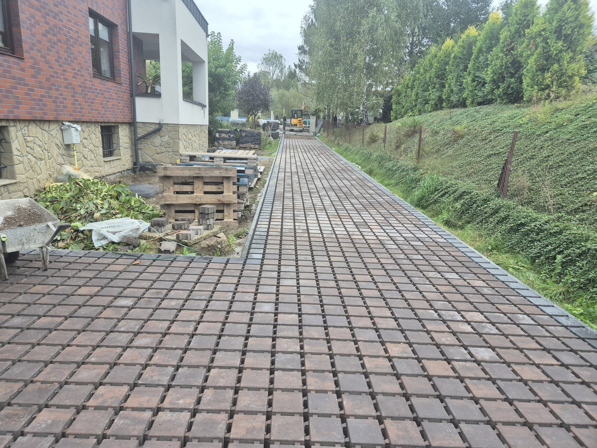 Układanie ażurowej kostki brukowej w Budzowie. Widok na długi podjazd z kostki, w tle dom i zieleń. Częściowo ułożona nawierzchnia, palety z kostką obok.