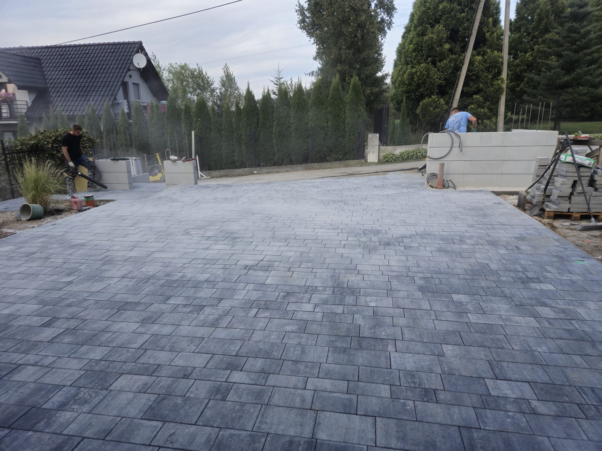 Ułożona szara kostka brukowa na posesji, widoczni dwaj pracownicy, jeden z dmuchawą, drugi przy betonowych elementach ogrodzenia. W tle dom i tuje.