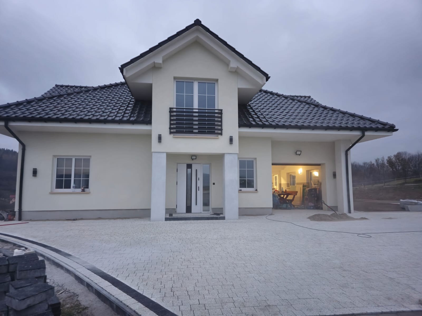 Nowoczesny dom jednorodzinny z jasną elewacją i ciemną dachówką. Ułożona kostka brukowa na podjeździe z elementami dekoracyjnymi. Widoczne prace wykończeniowe.