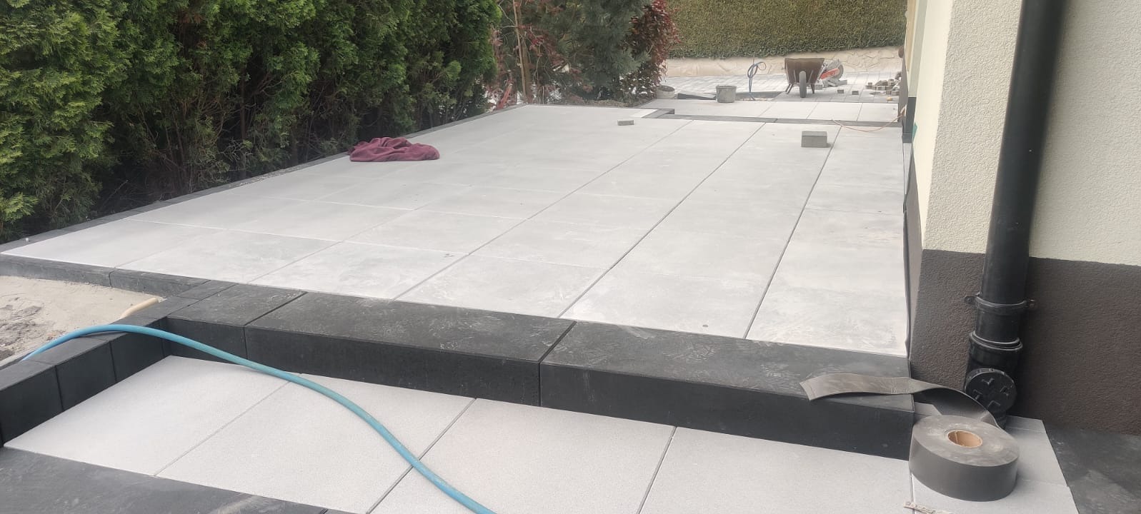 Jasnoszara kostka brukowa ułożona na tarasie, z ciemnymi obrzeżami. W tle zieleń i fragment ściany budynku z rurą spustową. Widoczne narzędzia i akcesoria brukarskie.