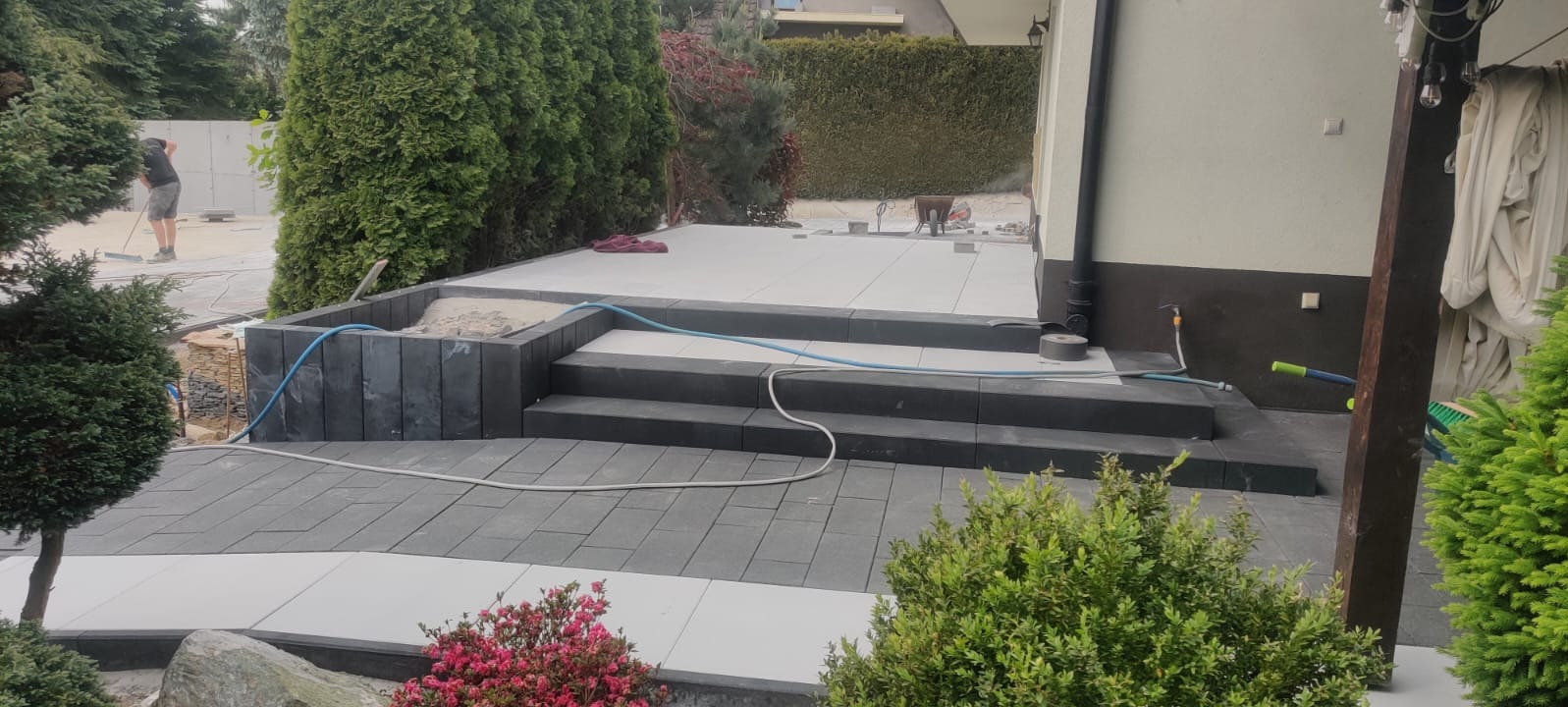 Nowoczesny taras z kostki brukowej w odcieniach szarości i bieli, z niskimi schodami i elementami małej architektury. W tle widoczna osoba podczas prac wykończeniowych.