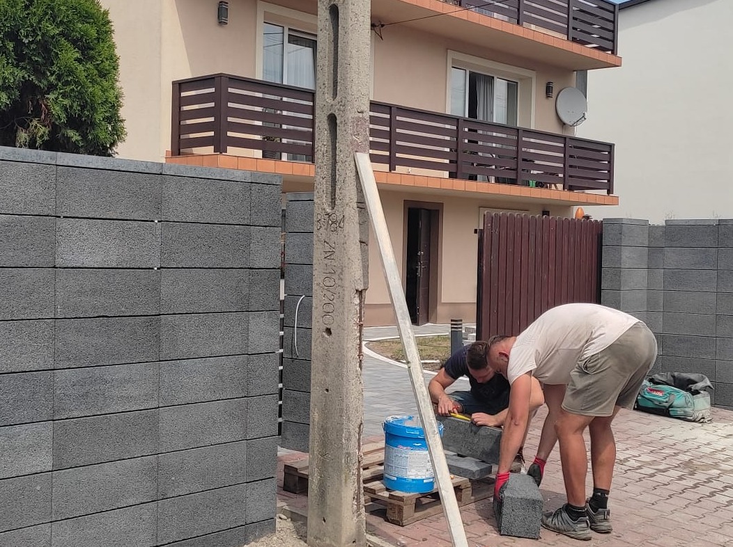 Budowa ogrodzenia z szarych bloczków betonowych. Dwóch mężczyzn przy pracy, jeden mierzy bloczek. W tle dom z balkonem i brązową bramą.