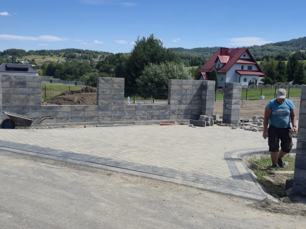 Budowa ogrodzenia z szarej kostki betonowej i układanie kostki brukowej na podjeździe, w tle dom z czerwonym dachem i zieleń. Widoczny pracownik w trakcie pracy.