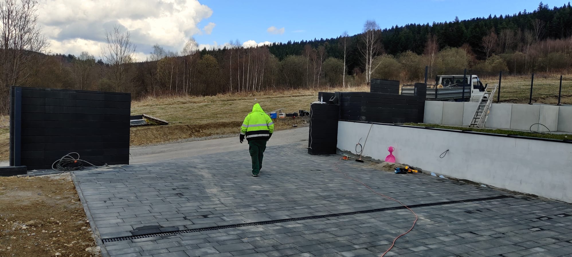 Ułożona szara kostka brukowa na podjeździe, widoczny fragment ogrodzenia z czarnych paneli, pracownik w odblaskowej kurtce. W tle zalesione wzgórza i błękitne niebo.